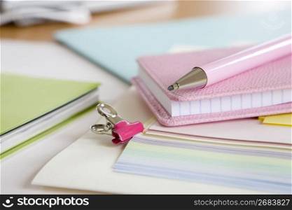 Pink clip hanging off a pile of pastel paper surrounded by notebooks
