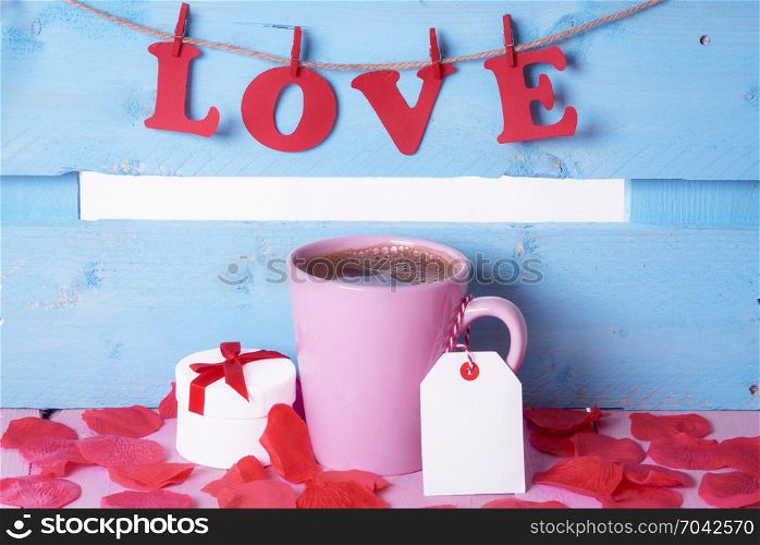 Pink cup of coffee with a label attached to its handle, near a gift, surrounded by soap rose petals and the word love written with red paper letters.