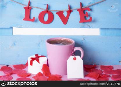 Pink cup of coffee with a label attached to its handle, near a gift, surrounded by soap rose petals and the word love written with red paper letters.