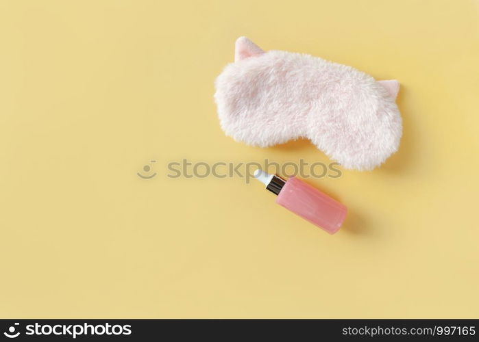 Pink fluffy fur sleep mask with small ears and bottle with serum for face on pastel yellow paper background. Top view. Concept of vivid dreams and skin care. Minimal style, copy space. Horizontal.