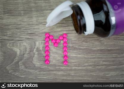 Pink pills forming shape to M alphabet on wood background&#xA;