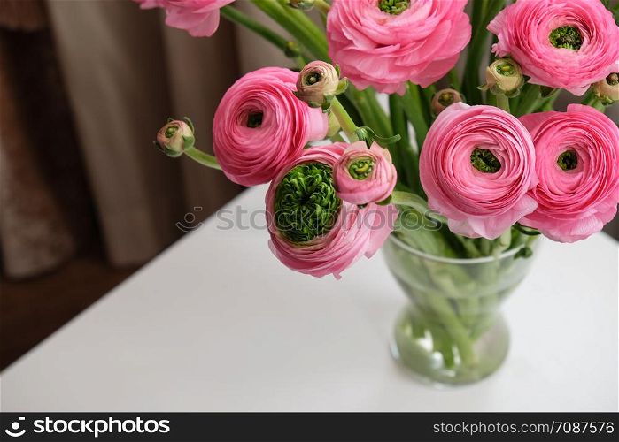 Pink Ranunculus bouquet in transparent glass vase on white table. Close-up. For flower delivery, social media. Soft selective focus. Copy space. Horizontal.