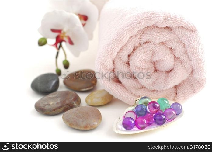 Pink rolled up towel with a stack of massage stones and bath beads on white background