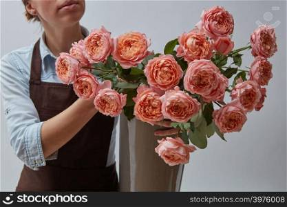 Pink roses in a vase. A female florist is holding a vase with flowers in hands around a gray background. The concept of a flower shop. Girl florist in a brown apron with a vase of pink roses on a gray background.
