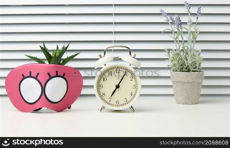 pink textile mask and round white alarm clock on the table, behind the white blinds