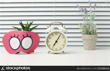 pink textile mask and round white alarm clock on the table, behind the white blinds