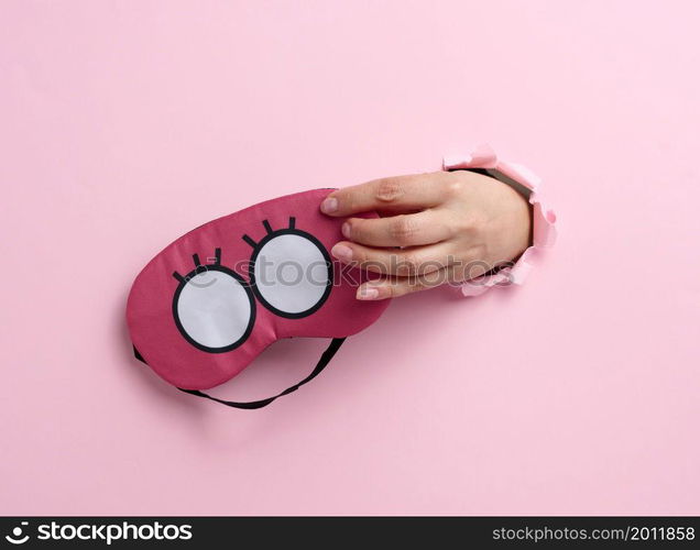 pink textile sleep mask in a female hand sticking out of a bull in a pink paper background, insomnia