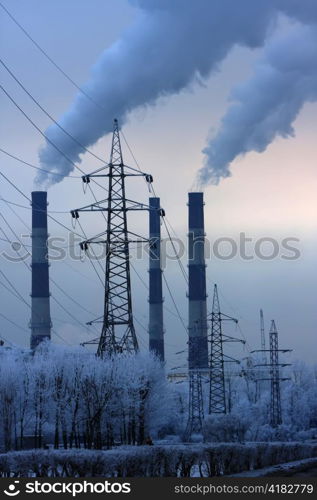 Pipe with smoke and line of electricity transmission on background of snow-covered city