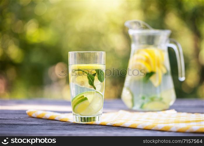 Pitcher and glass of homemade lemonade on a wooden table. Green natural background. The bright summer sun. Organic food concept. Pitcher and glass of homemade lemonade on wooden table