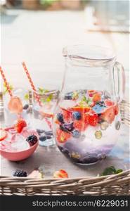 Pitcher with water, berries and ice cubes on table over garden terrace background. Home scene with summer drinks.