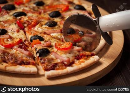 Pizza cutter wheel slicing ham pizza with capsicum and olives on wooden board on table. Ham pizza