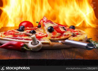 Pizza on wooden table with one slice on a lifter, real oven fire on backgroud