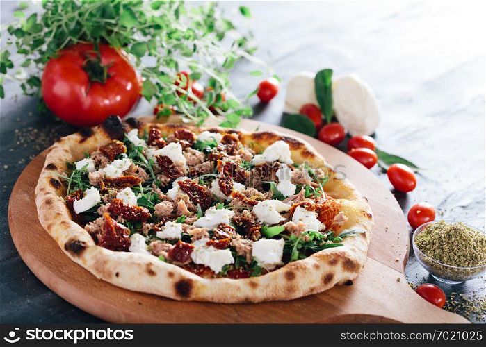 Pizza with dried tomatoes, mozzarella, sausage and rocket salad on wooden board. Oregano leaves, tomatoes and mozzarella in the background. Popular traditional food.. Pizza with dried tomatoes, mozzarella, sausage and rocket