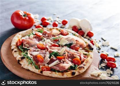 Pizza with ham, cheese, tomatoes and rocket salad on wooden board. Tomatoes, mozzarella and parmesan in the background. Popular traditional food.. Pizza with ham, cheese, tomatoes and rocket salad .