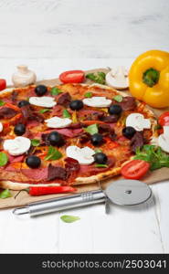 Pizza with ham, salami, tomato and chili pepper, mushrooms, on wooden table with pizza cutter