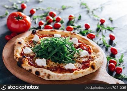 Pizza with salami, mozzarella and rocket salad on wooden board. Tomatoes and rocket salad leaves in the background. Popular traditional food.. Pizza with salami, mozzarella and rocket salad on wooden board.