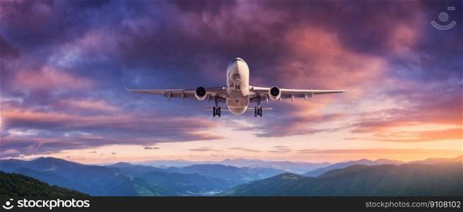Plane is flying in colorful sky at sunset. Landscape with passenger airplane, mountains, purple sky with orange and pink clouds. Aircraft is landing. Business travel. Aerial view. Transport 