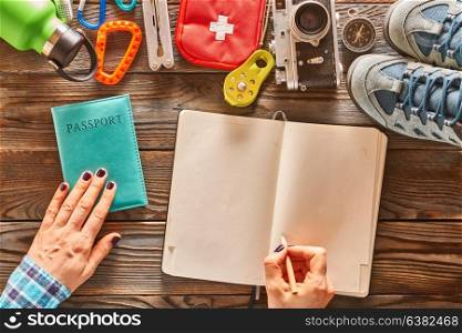 Planning a trip. Woman&rsquo;s hands with pencil over blank notebook. Travel items for hiking tourism still life over wooden background