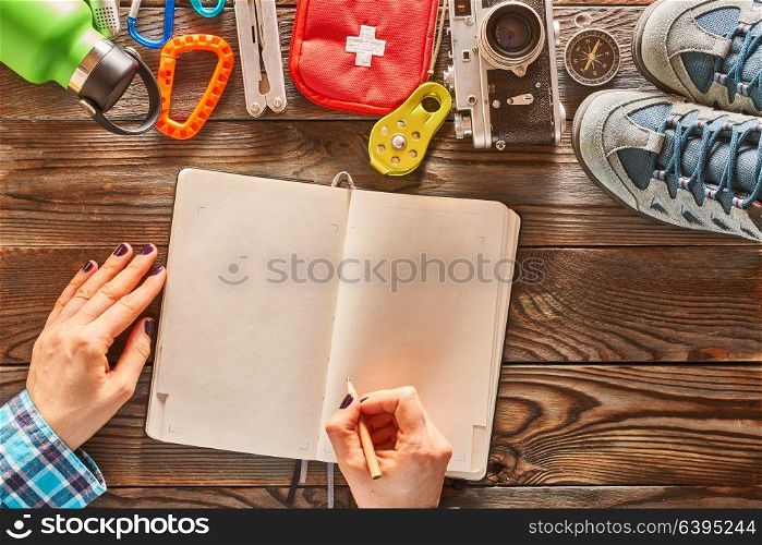 Planning a trip. Woman&rsquo;s hands with pencil over blank notebook. Travel items for hiking tourism still life over wooden background