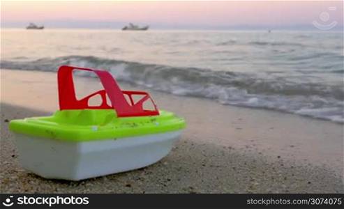 Plastic toy ship is standing on the sand in incoming sea waves at sunset. The wave is washing it away.