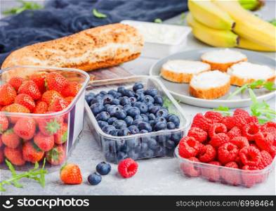 Plastic tray containers of fresh organic healthy beries and bread for fruit sandwiches. Blueberries, strawberries, bananas and raspberries on stone kitchen background.