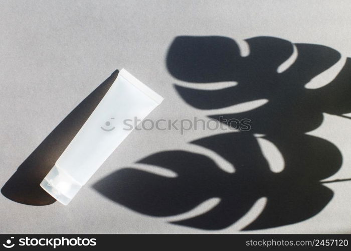 Plastic tube for cream or lotion on a gray background with a shadow of a montera leaves. Beauty concept.. Plastic tube for cream or lotion on a gray background with shadow of montera leaves. Beauty concept