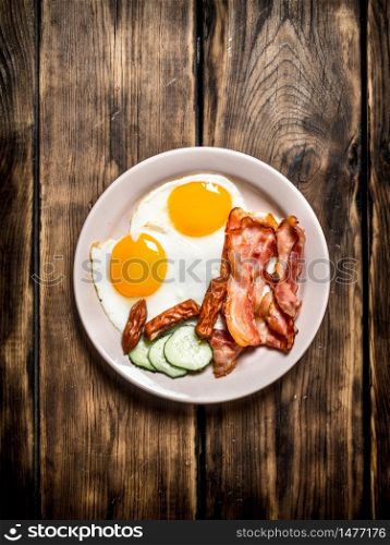 plate of fried eggs, bacon , cucumber and smoked sausages. On a wooden table.. plate of fried eggs, bacon , cucumber and smoked sausages.