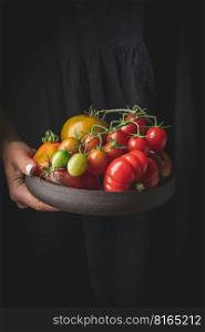 Plate of variety mix colorful tomatoes in woman hands. Fresh tomatoes on Rustic stile dark background.. Mix colorful tomatoes