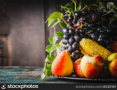 Plate with various harvesting fruits and vegetables: apple, corn, pear and grapes on rustic kitchen table at wooden background, side view
