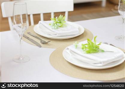 Plates with twigs of artificial flowers on beige round napkins, glasses and cutlery on a white table.Festive table setting for two persons.