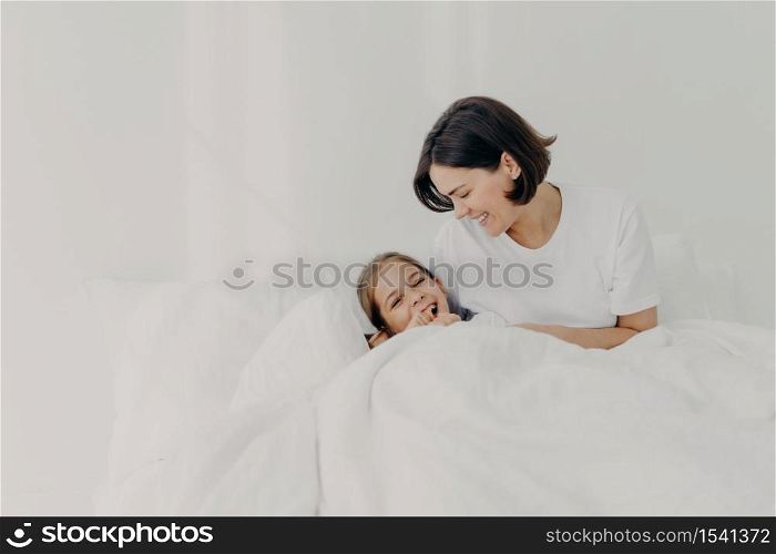 Playful caring mother and her daughter stay in bed for long time, pose under soft blanket, enjoy good morning, have happy smiles on faces, isolated over white background. Motherhood concept.