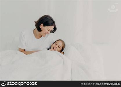Playful caring mother and her daughter stay in bed for long time, pose under soft blanket, enjoy good morning, have happy smiles on faces, isolated over white background. Motherhood concept.