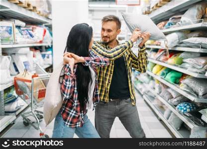 Playful couple in supermarket, pillow fight. Male and female customers on family shopping. Man and woman purchasing goods for the house. Playful couple in supermarket, pillow fight