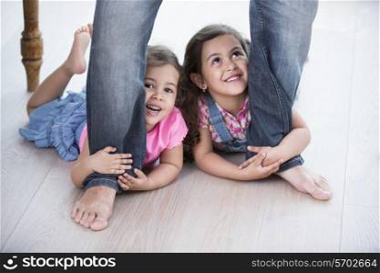 Playful girls holding father&rsquo;s legs on hardwood floor