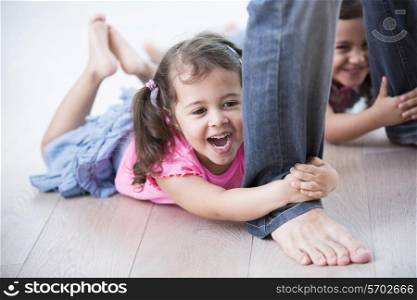 Playful girls holding father&rsquo;s legs on hardwood floor
