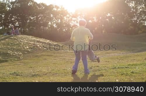 Playful grandma spinning her adorable toddler granson in circle in rays of setting sun in park. Cheerful grandmother playing with beloved grandchild in green field.