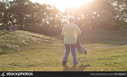 Playful grandma spinning her adorable toddler granson in circle in rays of setting sun in park. Cheerful grandmother playing with beloved grandchild in green field.