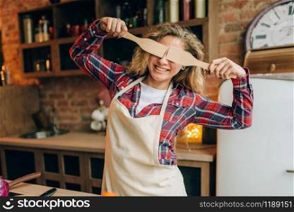 Playful housewife in an apron covers her eyes with wooden spatulas, kitchen interior on background. Happy female cook making healthy vegetarian food, salad cooking. Housewife covers her eyes with wooden spatulas