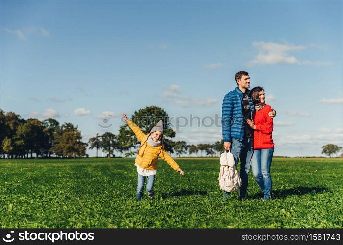 Playful little child runs on green meadow, plays near parents, who embrace each other, look into distance with thoughtful expressions. Family has walk or stroll in countryside, rest after noise city