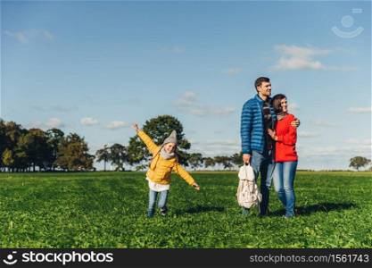 Playful little child runs on green meadow, plays near parents, who embrace each other, look into distance with thoughtful expressions. Family has walk or stroll in countryside, rest after noise city