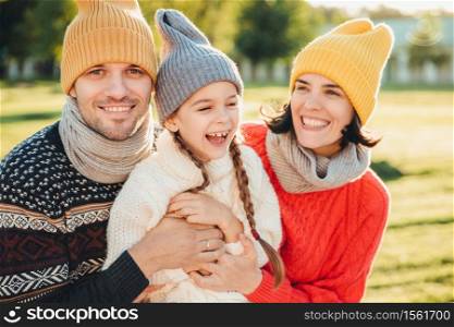 Playful little child with pigtails wears warm clothes, spends free time with lovely affectionate parents, have happy expression, feel relaxed. Three young family members enjoy pleasant moments