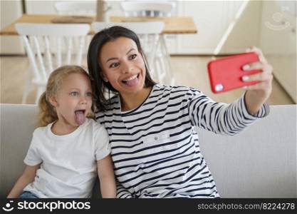 Playful mom and little child daughter holding phone, taking selfie photo, showing tongues together, recording funny video for social network. Small girl and mother having fun sitting on couch at home.. Mom, little daughter take selfie photo by phone, show tongues together, having fun, sitting on sofa
