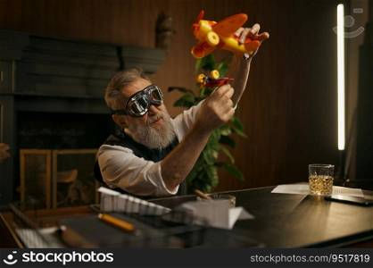 Playful senior boss playing toy plane at work table. Mature grey-haired businessman wearing elegant clothes and aircraft pilot goggles having fun and relax. Playful senior boss or businessman playing toy plane at work table