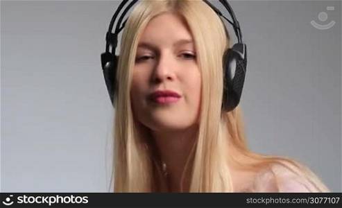 Playful teenage girl with headset listening to the music and swaying groovily to the beat on white background. Sexy blonde woman flirting with camera, seductively putting her finger on red lips and sending air kiss while enjoying music in headphones.