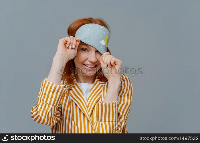 Playful woman with ginger hair looks from sleep mask, wears striped pajamas, has cheerful expression, poses over grey background, enjoys sleeping at home. Healthy sleep and relaxation concept