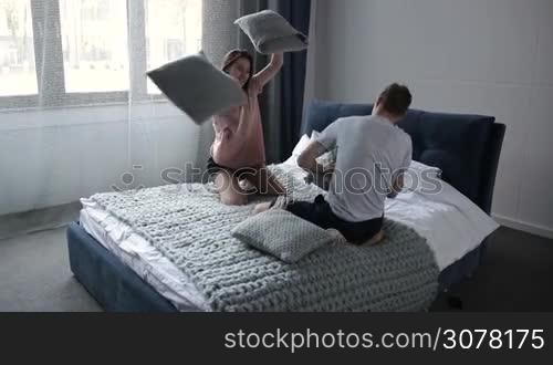 Playful young attractive couple in pajamas fooling around and having pillow figth in bedroom. Cheerful hipster family spending leisure together in bed, fighting with pillows. Slow motion. Steadicam stabilized shot.