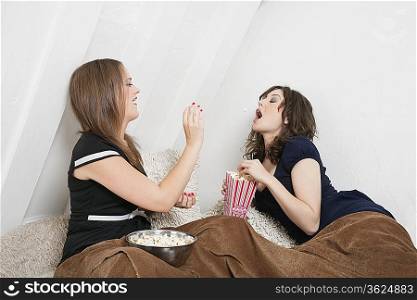 Playful young female throwing popcorn into friend&acute;s mouth in bed