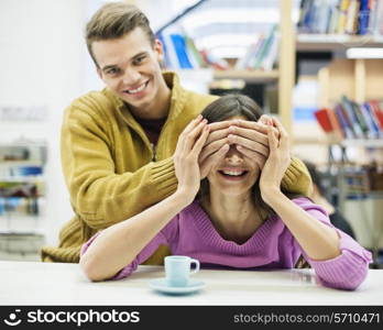 Playful young man covering woman&rsquo;s eyes in library