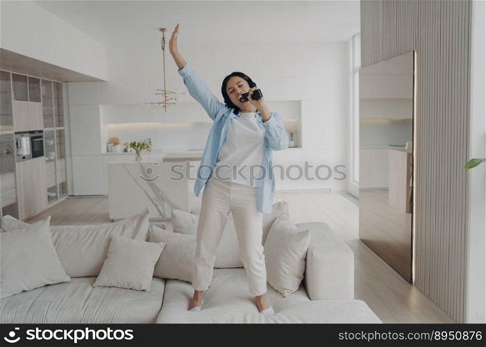 Playful young woman is listening to music in earphones, singing in microphone. Excited arab girl in headphones dancing on sofa in morning. Positive emotions, relaxation and entertainment concept.. Playful young woman is listening music in earphones, singing in microphone, dancing on sofa.