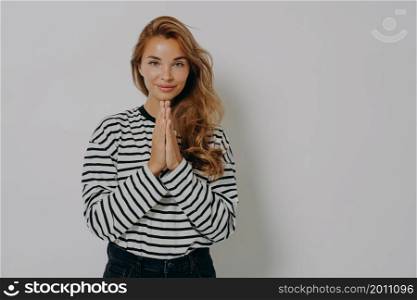Pleading young woman in casual striped shirt holding hands folded in praying gesture, asking for help, pressing palms together while posing isolated on grey studio background with copy space for text. Pleading young blonde woman girl in casual striped shirt holding hands folded in praying gesture